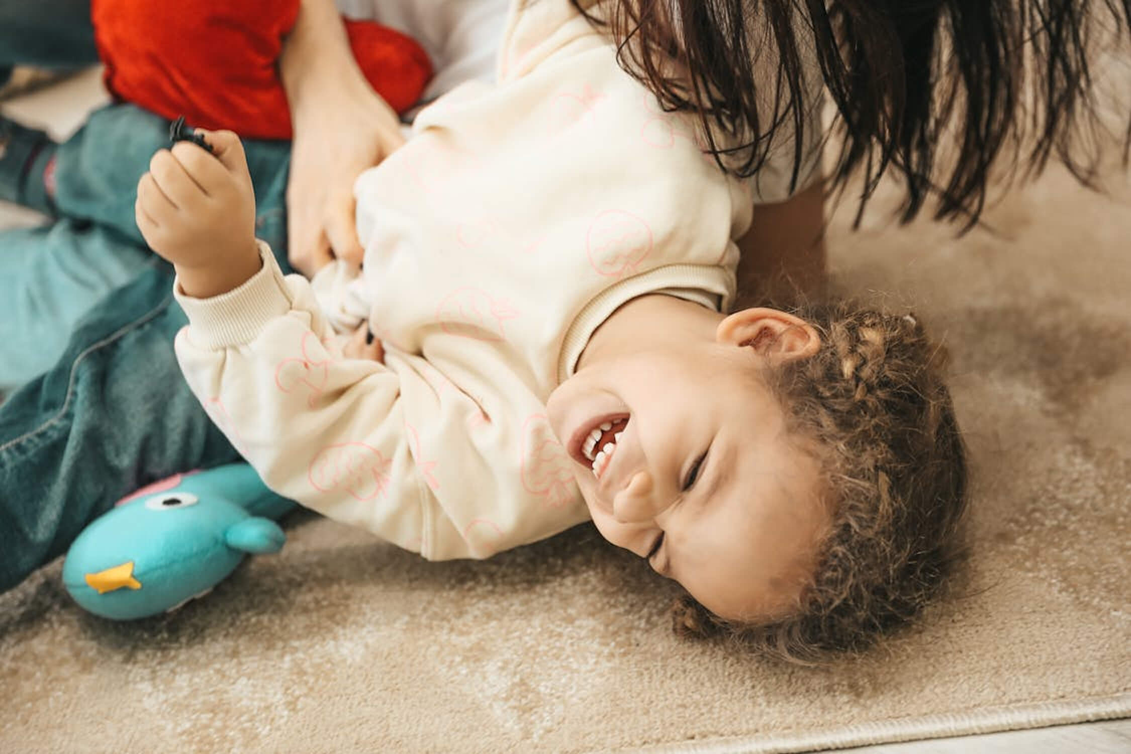 A young child laughs joyfully while lying on a carpeted floor, with a stuffed toy bird nearby and an adult partially visible, playing with them.