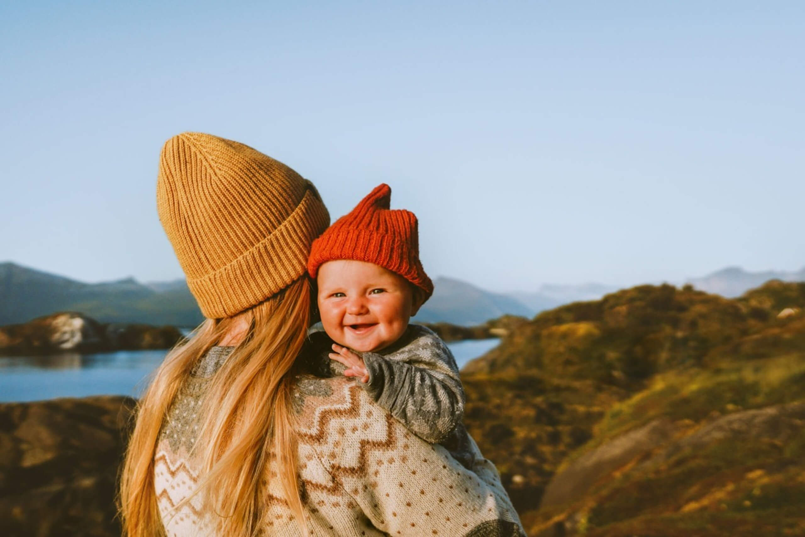 A woman with long blonde hair wearing a tan knit hat holds a smiling baby in a red knit hat, outdoors near a lake and mountains under a clear blue sky.
