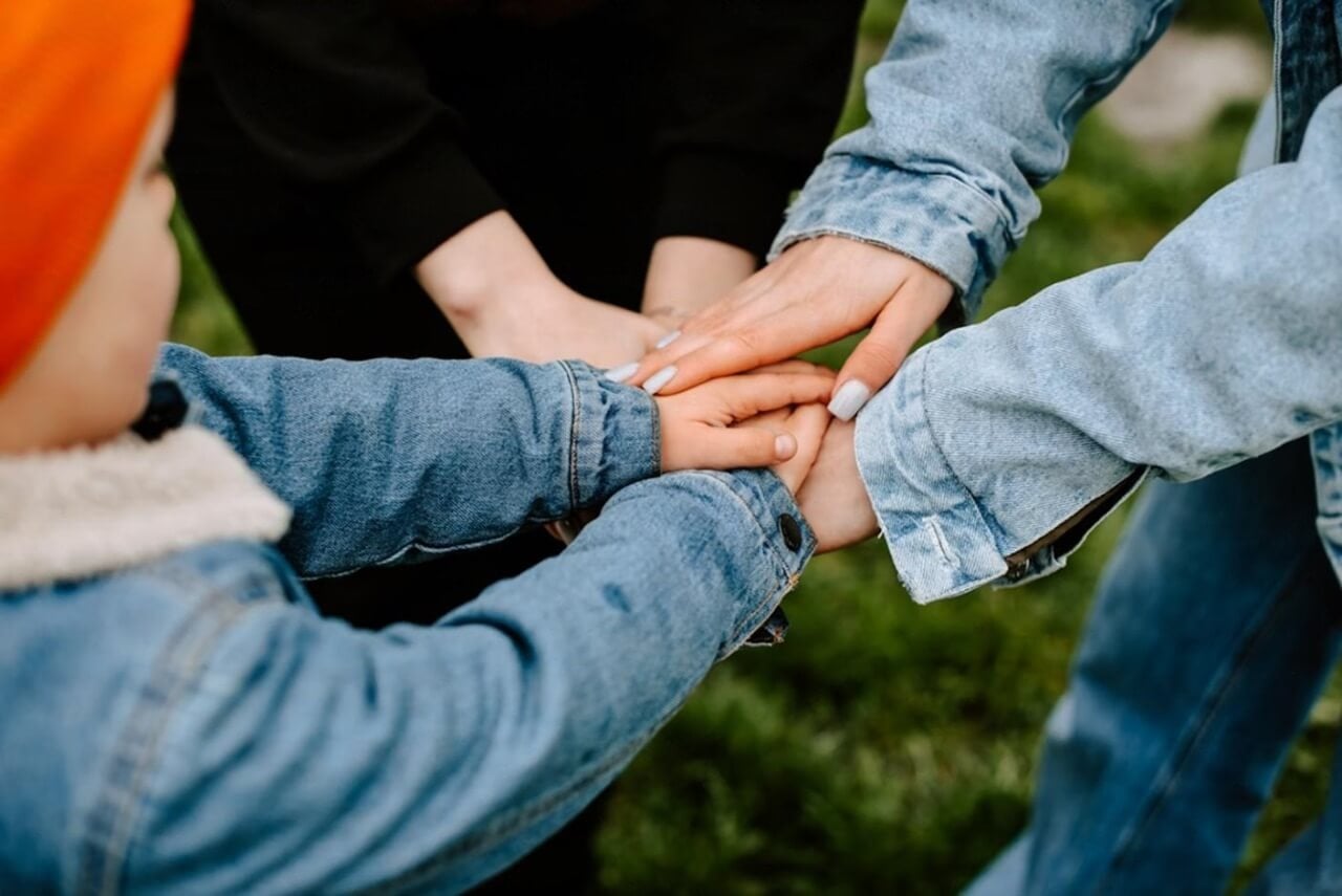 Lähikuva kolmesta ihmisestä, jotka pinoavat käsiään yhteen ulkona; yhdellä lapsella on oranssi hattu ja sininen takki ja kahdella aikuisella on farkut ja takit, ja he seisovat vihreällä nurmikolla.